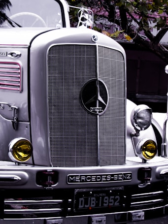 A classic Mercedes-Benz truck with a prominent front grille and headlights, bearing a vintage style. The emblem and the word 'DIESEL' are clearly visible on the grille. Yellow-tinted round headlights add to the retro aesthetic. The license plate reads 'DJB 1952', and the silver and chrome finish reflects the surrounding environment with hints of matte green leaves above.