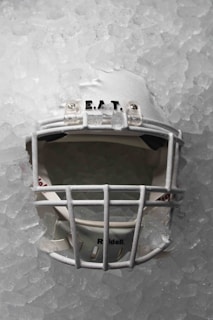 A white football helmet embedded in a layer of ice, with the faceguard clearly visible. The surface of the ice is uneven and textured, and the helmet appears slightly frosted, emphasizing its cold environment.