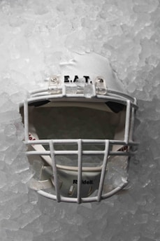 A white football helmet embedded in a layer of ice, with the faceguard clearly visible. The surface of the ice is uneven and textured, and the helmet appears slightly frosted, emphasizing its cold environment.