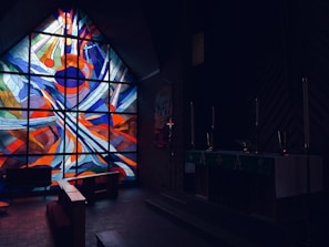 A dimly lit church interior with a vibrant stained glass window portraying abstract patterns in bright red, blue, and orange colors. The darkened room contains pews and an altar with several tall candle holders, evoking a solemn atmosphere.