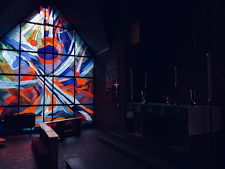 A dimly lit church interior with a vibrant stained glass window portraying abstract patterns in bright red, blue, and orange colors. The darkened room contains pews and an altar with several tall candle holders, evoking a solemn atmosphere.