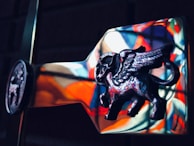 A detailed and artistic depiction of a flying mythological creature with wings, resembling a bull or a similar animal, crafted in a metallic material. The background contains colorful reflections, possibly from a stained glass window, creating a vibrant backdrop.