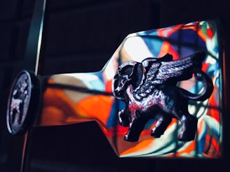 A detailed and artistic depiction of a flying mythological creature with wings, resembling a bull or a similar animal, crafted in a metallic material. The background contains colorful reflections, possibly from a stained glass window, creating a vibrant backdrop.