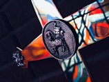 A close-up of a metallic cross with intricate designs, including a detailed engraving of a lamb carrying a flag at the center. The background features colorful stained glass patterns, casting vibrant reflections on the cross.