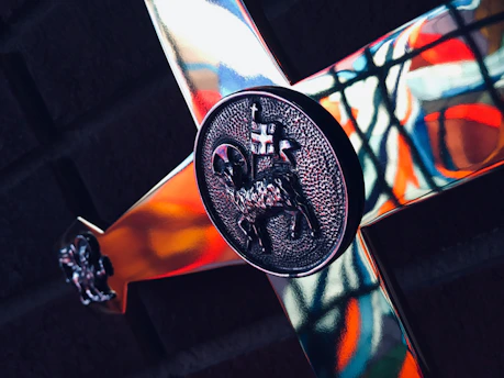 A close-up of a metallic cross with intricate designs, including a detailed engraving of a lamb carrying a flag at the center. The background features colorful stained glass patterns, casting vibrant reflections on the cross.