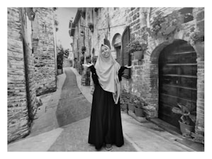 A person wearing a modest outfit stands in a narrow, cobblestone street lined with stone buildings. Potted plants are visible along the walls, and wooden shutters decorate the windows. The scene appears to be set in a quaint, historic European town.