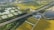 Aerial view of a rural landscape featuring a network of fields with green and yellow crops, intersected by a highway. Several bodies of water are scattered throughout the scenery, bordered by clusters of trees. There is also a group of industrial buildings situated near the road.