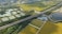 Aerial view of a rural landscape featuring a network of fields with green and yellow crops, intersected by a highway. Several bodies of water are scattered throughout the scenery, bordered by clusters of trees. There is also a group of industrial buildings situated near the road.