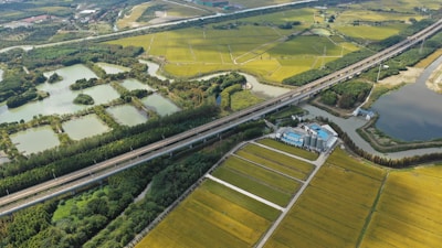 Aerial view of a rural landscape featuring a network of fields with green and yellow crops, intersected by a highway. Several bodies of water are scattered throughout the scenery, bordered by clusters of trees. There is also a group of industrial buildings situated near the road.