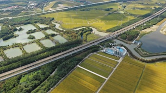 Aerial view of a rural landscape featuring a network of fields with green and yellow crops, intersected by a highway. Several bodies of water are scattered throughout the scenery, bordered by clusters of trees. There is also a group of industrial buildings situated near the road.