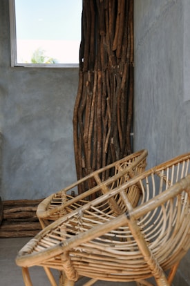 Rattan chairs with an open, woven design are placed against a concrete wall. A decorative wooden panel made of vertical logs adds a rustic element. Natural light filters through a small window, creating a serene and earthy ambiance.