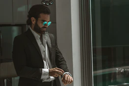 Stylish man adjusting his cufflinks in front of a dark mirror reflecting subtle blue lighting.