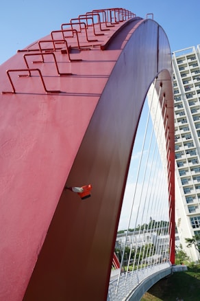 A large red arch structure features prominently, with geometrical guardrails on top. A red and orange security camera is attached to the side. In the background, there is a modern high-rise building and a cable-stayed bridge with white suspension cables extending across a span. The sky is clear and blue, contributing to the overall detail of the environment.