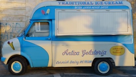 A vintage ice cream van painted blue and cream, featuring colorful ice cream illustrations on the sides. Writing on the van includes phrases like 'Traditional Ice Cream,' 'Antica Gelateria,' and 'Wholesome and Delicious.' The van is parked against a graffiti-covered wall.