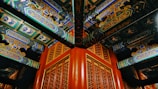 Intricately painted wooden beams and decorative patterns in a traditional architectural style dominate the scene. The vibrant colors and detailed motifs showcase artistic craftsmanship, with each beam featuring intricate designs and script-like symbols. The wooden grid pattern below complements the elaborate ceiling.