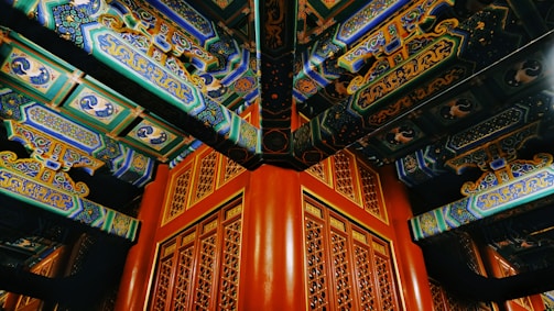 Intricately painted wooden beams and decorative patterns in a traditional architectural style dominate the scene. The vibrant colors and detailed motifs showcase artistic craftsmanship, with each beam featuring intricate designs and script-like symbols. The wooden grid pattern below complements the elaborate ceiling.