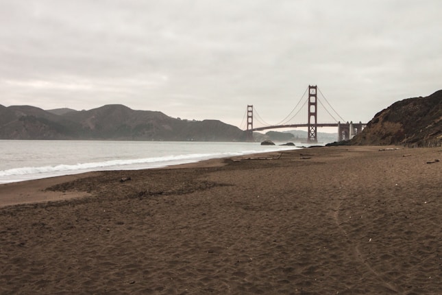 A sandy beach stretches along the coastline with gentle waves lapping at the shore. In the background, the iconic Golden Gate Bridge spans across the bay under a cloudy sky. Hills and rocky formations can be seen in the distance.