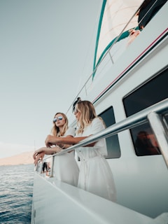 Two women wearing white outfits and sunglasses are leaning on the railing of a large yacht. The sky is clear, and the ocean is calm, suggesting a sunny day. The yacht appears modern with a clean, white exterior. One of the women has long blonde hair, and they are both gazing at the horizon, giving a sense of relaxation and enjoyment.