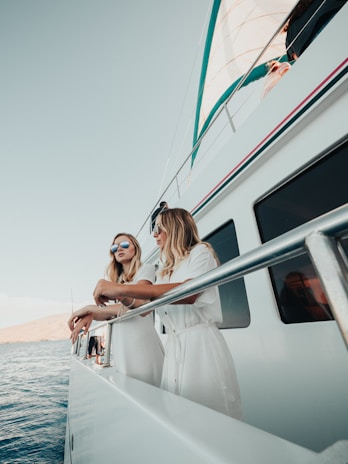 Two women wearing white outfits and sunglasses are leaning on the railing of a large yacht. The sky is clear, and the ocean is calm, suggesting a sunny day. The yacht appears modern with a clean, white exterior. One of the women has long blonde hair, and they are both gazing at the horizon, giving a sense of relaxation and enjoyment.
