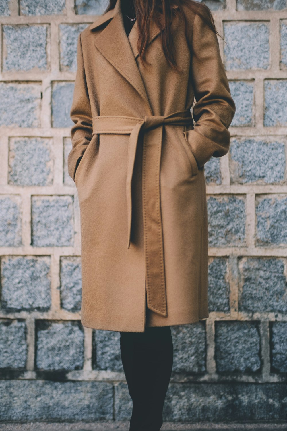 donna che indossa un cappotto marrone monopetto in piedi davanti a un muro di mattoni grigi