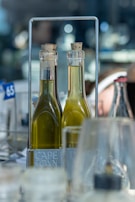 Two clear glass bottles with cork stoppers are filled with a yellow liquid, likely oil or vinegar. They are placed in a metal holder labeled 'CAPE TOWN'. The background is blurred, featuring indistinct shapes and colors, suggesting a restaurant or kitchen setting with a bokeh effect.