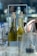 Two clear glass bottles with cork stoppers are filled with a yellow liquid, likely oil or vinegar. They are placed in a metal holder labeled 'CAPE TOWN'. The background is blurred, featuring indistinct shapes and colors, suggesting a restaurant or kitchen setting with a bokeh effect.