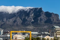 A prominent mountain with a flat top dominates the background, partially covered by a layer of fluffy white clouds. Below the mountain, there is an urban landscape with modern buildings and a yellow frame structure labeled 'Table Mountain, Cape Town, South Africa.' The sky is clear and blue, contrasting with the rugged texture of the mountain.