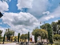 A peaceful park setting with manicured trees and vintage-style lampposts under a mostly blue sky with a few fluffy white clouds. In the background, colorful buildings with red, yellow, and white hues add a vibrant touch.