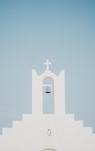 A minimalist white church facade with a prominent bell hanging under an archway, topped by a cross. The background is a clear, pale blue sky.