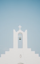 A minimalist white church facade with a prominent bell hanging under an archway, topped by a cross. The background is a clear, pale blue sky.