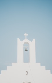 A minimalist white church facade with a prominent bell hanging under an archway, topped by a cross. The background is a clear, pale blue sky.
