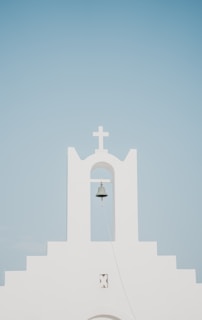 A minimalist white church facade with a prominent bell hanging under an archway, topped by a cross. The background is a clear, pale blue sky.