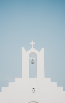 A minimalist white church facade with a prominent bell hanging under an archway, topped by a cross. The background is a clear, pale blue sky.