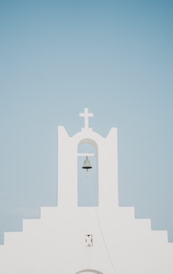 A minimalist white church facade with a prominent bell hanging under an archway, topped by a cross. The background is a clear, pale blue sky.