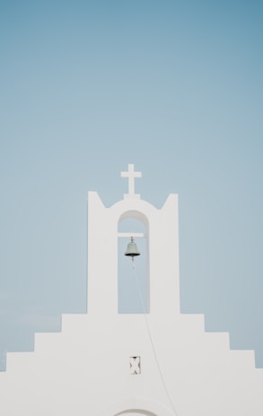 A minimalist white church facade with a prominent bell hanging under an archway, topped by a cross. The background is a clear, pale blue sky.