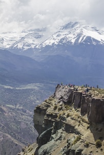 A group of people standing at the edge of a rocky cliff, overlooking a vast mountainous landscape with snow-capped peaks under a cloudy sky. The foreground displays rugged terrain with sparse vegetation, while the distant mountains create a dramatic backdrop.