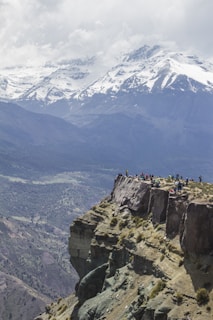 A group of people standing at the edge of a rocky cliff, overlooking a vast mountainous landscape with snow-capped peaks under a cloudy sky. The foreground displays rugged terrain with sparse vegetation, while the distant mountains create a dramatic backdrop.