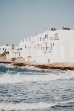 White buildings with blue accents are situated along a coastal area. Waves crash against the rocky shore, creating a scenic and serene atmosphere. The buildings have a Mediterranean architectural style, and there are some visible solar panels on the rooftops.