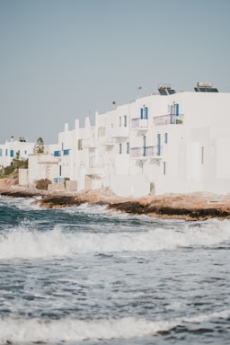 White buildings with blue accents are situated along a coastal area. Waves crash against the rocky shore, creating a scenic and serene atmosphere. The buildings have a Mediterranean architectural style, and there are some visible solar panels on the rooftops.