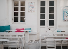 A charming outdoor seating area featuring white furniture against a backdrop of white plaster walls. Vibrant teal and pink cushions add a pop of color to the benches. Two doors and a window are visible, along with decorative signs that suggest a relaxed, casual atmosphere. The setting is reminiscent of a cozy café or a seaside restaurant.