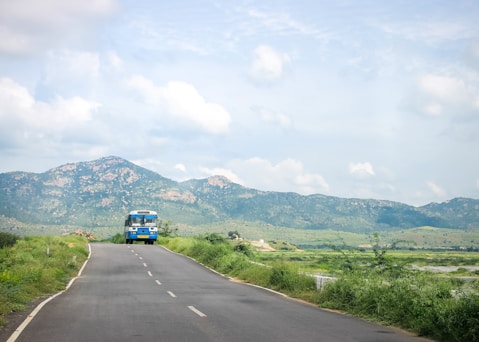 A solitary bus travels along a winding road surrounded by lush greenery. In the background, majestic mountains rise against a partly cloudy sky, creating a scenic and tranquil landscape.