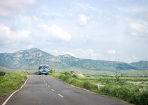 A solitary bus travels along a winding road surrounded by lush greenery. In the background, majestic mountains rise against a partly cloudy sky, creating a scenic and tranquil landscape.
