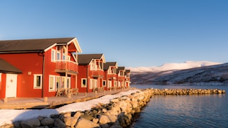 A row of red wooden houses with white trim line a snowy shore beside a calm body of water. The houses feature decks and small balconies, facing a landscape of snow-covered mountains in the background under a clear blue sky.