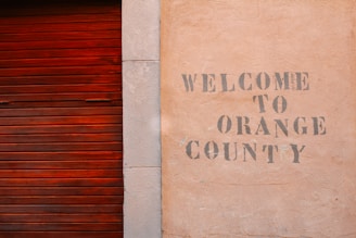 A textured wall with the words 'Welcome to Orange County' stenciled in a faded, rustic font next to a wooden, reddish-brown slatted door.