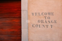 A textured wall with the words 'Welcome to Orange County' stenciled in a faded, rustic font next to a wooden, reddish-brown slatted door.