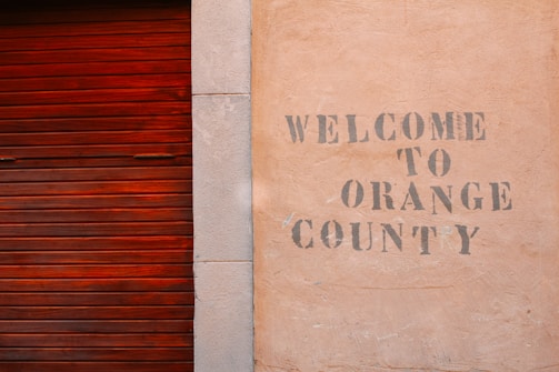 A textured wall with the words 'Welcome to Orange County' stenciled in a faded, rustic font next to a wooden, reddish-brown slatted door.