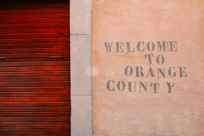 A textured wall with the words 'Welcome to Orange County' stenciled in a faded, rustic font next to a wooden, reddish-brown slatted door.