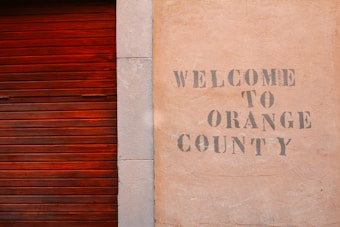 A textured wall with the words 'Welcome to Orange County' stenciled in a faded, rustic font next to a wooden, reddish-brown slatted door.