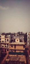 A residential area features multi-story buildings in an urban setting. Some buildings have balconies with decorative railings and rooftops adorned with antennas and satellite dishes. The scene is under a vast sky with a flock of birds flying in the distance. In the backdrop, distant trees and a hazy skyline are visible.
