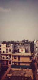 A residential area features multi-story buildings in an urban setting. Some buildings have balconies with decorative railings and rooftops adorned with antennas and satellite dishes. The scene is under a vast sky with a flock of birds flying in the distance. In the backdrop, distant trees and a hazy skyline are visible.